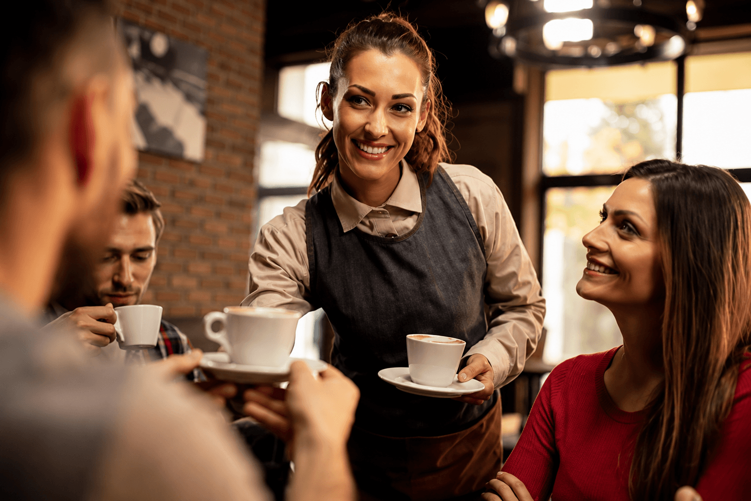 A barista smiling as she delivers cappuccinos at a wooden cafe table.
