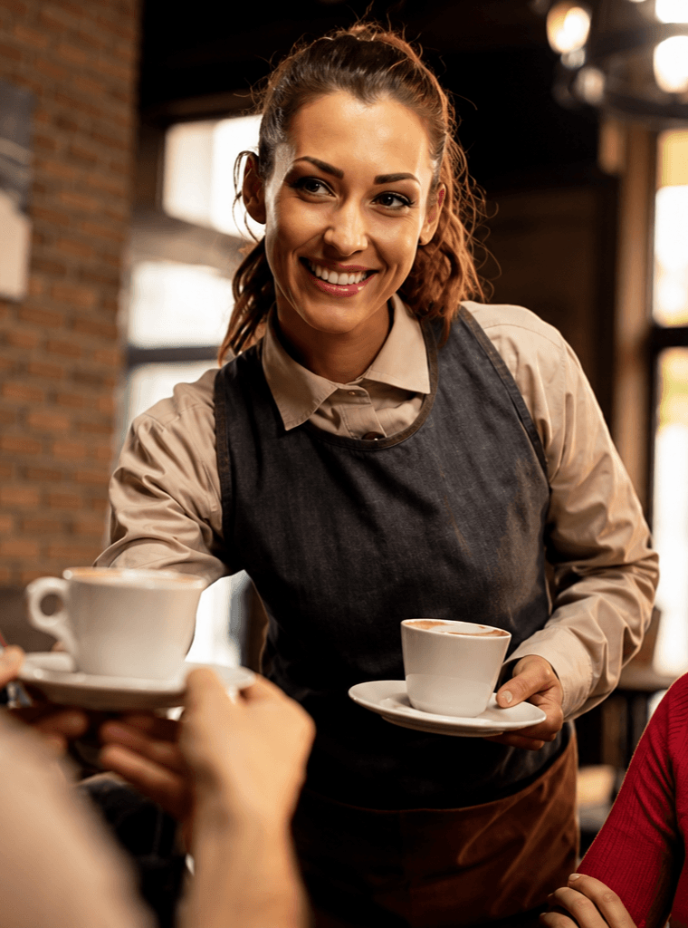 A barista smiling as she delivers cappuccinos at a wooden cafe table.